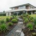 Maison individuelle implantée sur un terrain en pente, avec une noue végétalisée et une tranchée drainante recueillant les eaux de pluie dans un jardin humide après une averse.