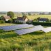 Vue réaliste d’un parc agrivoltaïque installé en lisière d’un village français, avec des panneaux solaires espacés au-dessus de prairies, des maisons rurales et des haies bocagères.